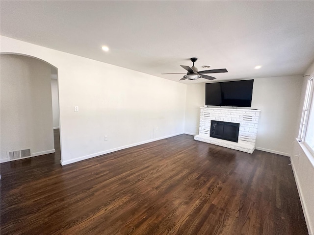 unfurnished living room featuring a ceiling fan, dark wood-type flooring, a stone fireplace, baseboards, and arched walkways