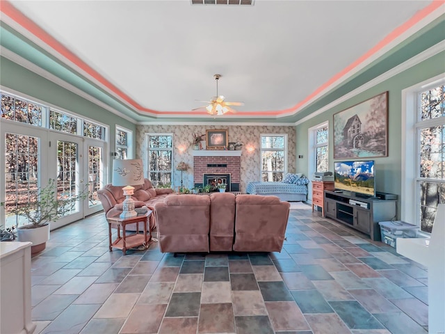 living area featuring a ceiling fan, healthy amount of natural light, a tray ceiling, a brick fireplace, and ornamental molding