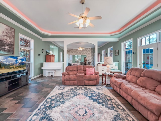 living area featuring ceiling fan, a tray ceiling, arched walkways, crown molding, and baseboards
