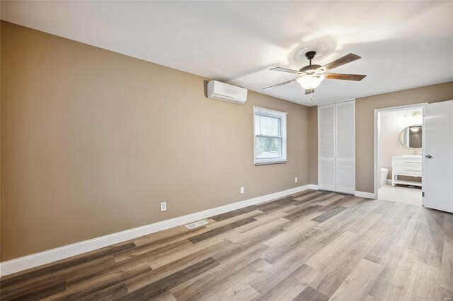 unfurnished bedroom with a wall mounted AC, light wood-style floors, ensuite bathroom, baseboards, and ceiling fan
