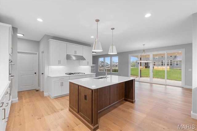 kitchen with a sink, under cabinet range hood, stainless steel gas cooktop, light countertops, and a center island with sink
