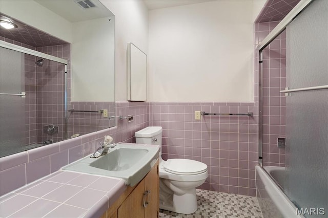 bathroom featuring toilet, a wainscoted wall, bath / shower combo with glass door, vanity, and tile walls