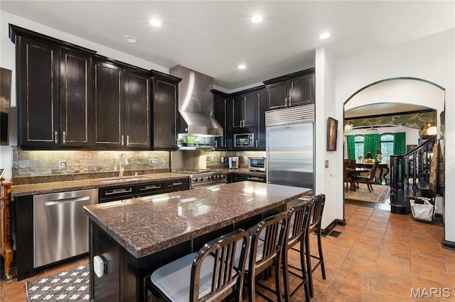 kitchen with built in appliances, a sink, wall chimney range hood, backsplash, and a breakfast bar area