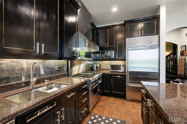 kitchen featuring premium appliances, a sink, arched walkways, tasteful backsplash, and dark stone countertops