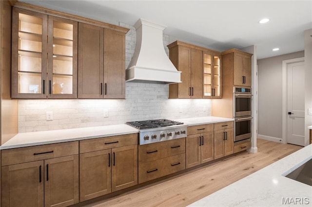 19 Beaver Dr, Creve Coeur, MO 63141 - kitchen featuring custom range hood, appliances with stainless steel finishes, light wood-type flooring, decorative backsplash, and glass insert cabinets