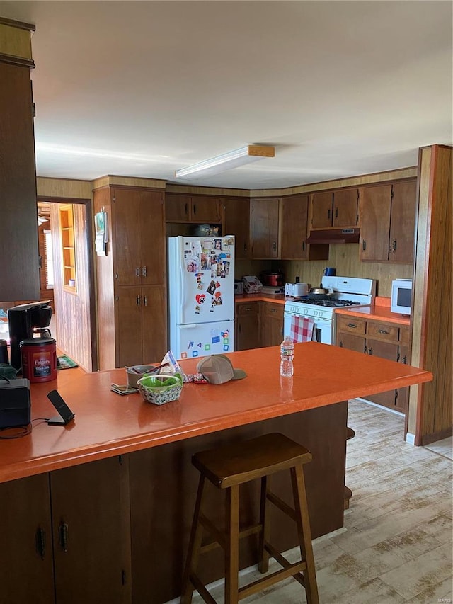 kitchen with white appliances, under cabinet range hood, a peninsula, light countertops, and brown cabinets