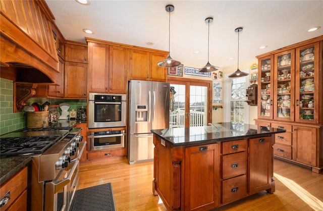 kitchen featuring appliances with stainless steel finishes, light wood-type flooring, brown cabinetry, glass insert cabinets, and tasteful backsplash