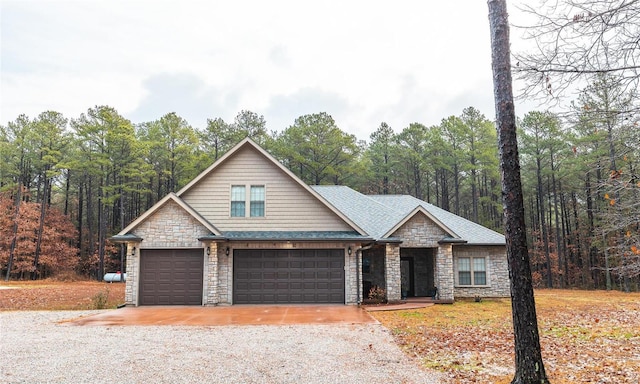 craftsman-style house with stone siding, gravel driveway, a shingled roof, and a forest view