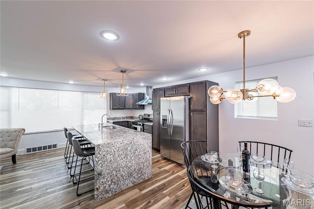 1101 W Parkedge Ln, Saint Louis, MO 63130 - kitchen with appliances with stainless steel finishes, wall chimney range hood, a kitchen breakfast bar, wood finished floors, and dark brown cabinetry