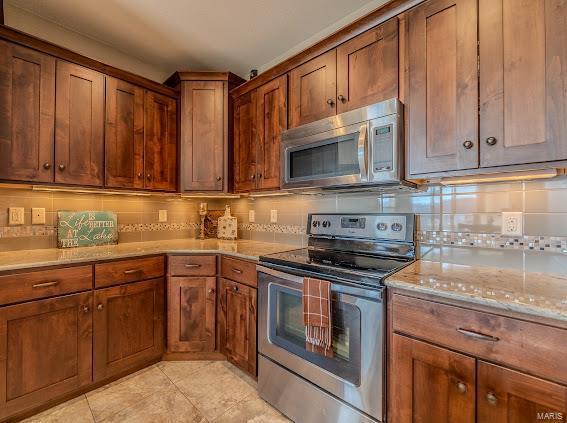 kitchen featuring stainless steel appliances, tasteful backsplash, light tile patterned floors, light stone counters, and brown cabinetry