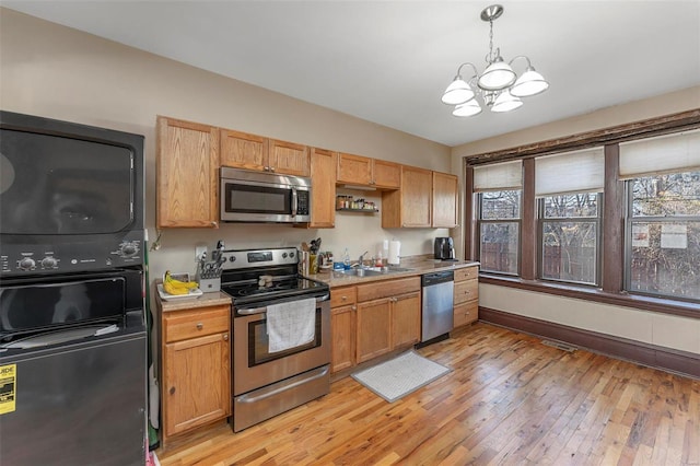 kitchen featuring appliances with stainless steel finishes, stacked washing machine and dryer, a chandelier, a sink, and light wood-style flooring