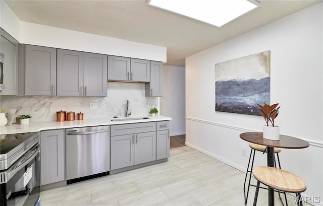 kitchen with stainless steel appliances, gray cabinetry, a sink, light countertops, and backsplash