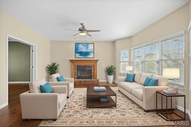 living room featuring baseboards, wood finished floors, a ceiling fan, and a brick fireplace
