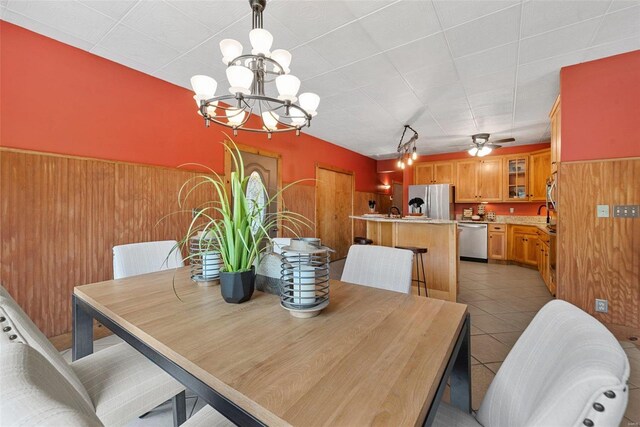 dining area with a chandelier, wooden walls, a ceiling fan, light tile patterned floors, and a wainscoted wall