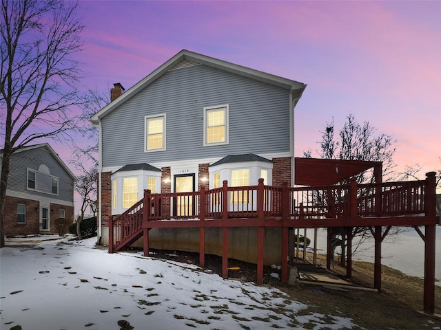 snow covered rear of property featuring brick siding, a wooden deck, a chimney, and stairs