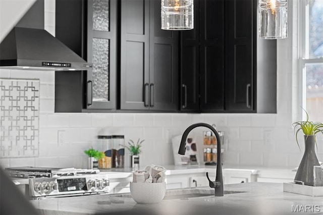 kitchen featuring wall chimney range hood, dark cabinets, tasteful backsplash, and light stone countertops