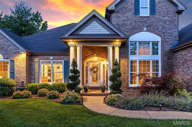 doorway to property featuring brick siding, a yard, and a shingled roof