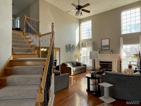 living room featuring a ceiling fan, stairs, a towering ceiling, wood finished floors, and a glass covered fireplace