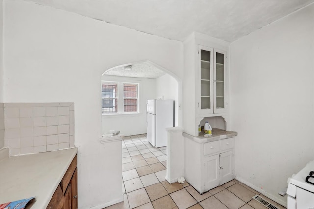 kitchen with freestanding refrigerator, white cabinets, light tile patterned floors, arched walkways, and glass insert cabinets