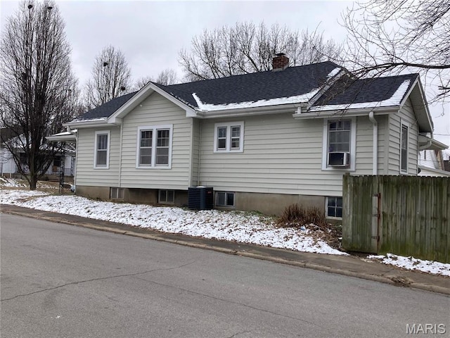 view of snowy exterior with a chimney, cooling unit, roof with shingles, and fence