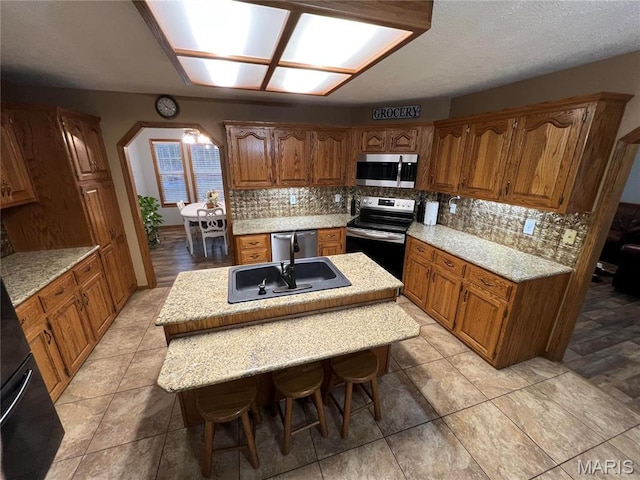 kitchen featuring appliances with stainless steel finishes, a sink, a center island with sink, decorative backsplash, and brown cabinetry