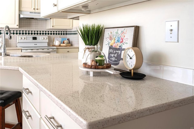 kitchen view of under cabinet range hood, electric stove, decorative backsplash, light stone countertops, and a sink
