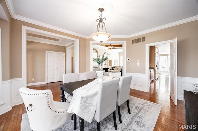 dining space featuring dark wood finished floors, a wainscoted wall, crown molding, and a ceiling fan