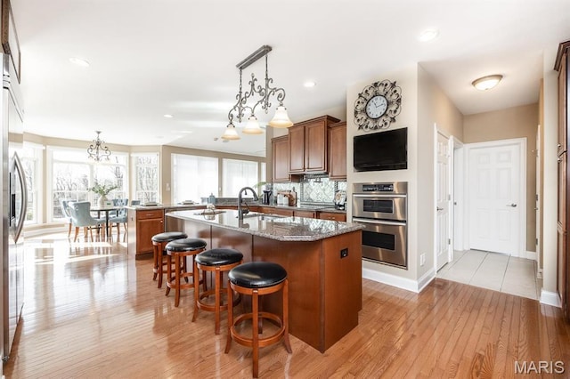 kitchen featuring stainless steel appliances, a center island with sink, a sink, brown cabinets, and light wood-style flooring