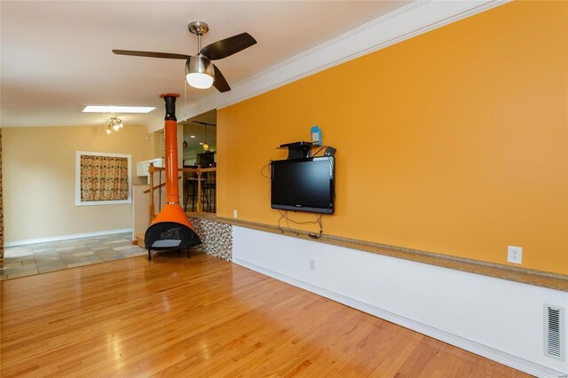 living area with a wood stove, ceiling fan, wood finished floors, baseboards, and ornamental molding