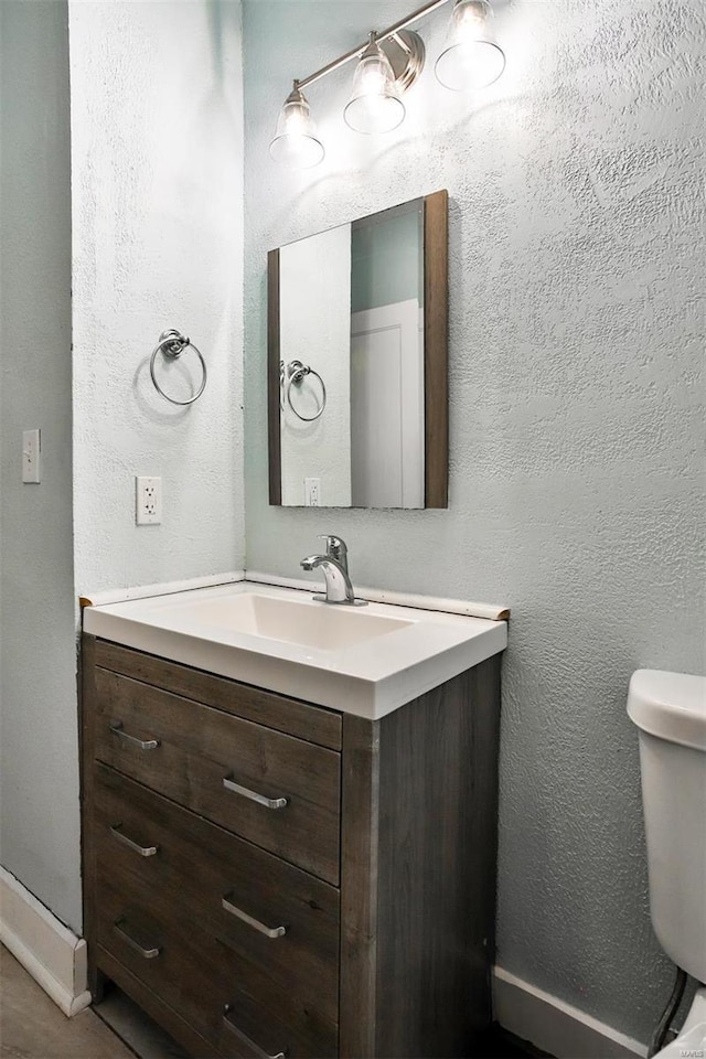 half bath featuring vanity, baseboards, a textured wall, and toilet