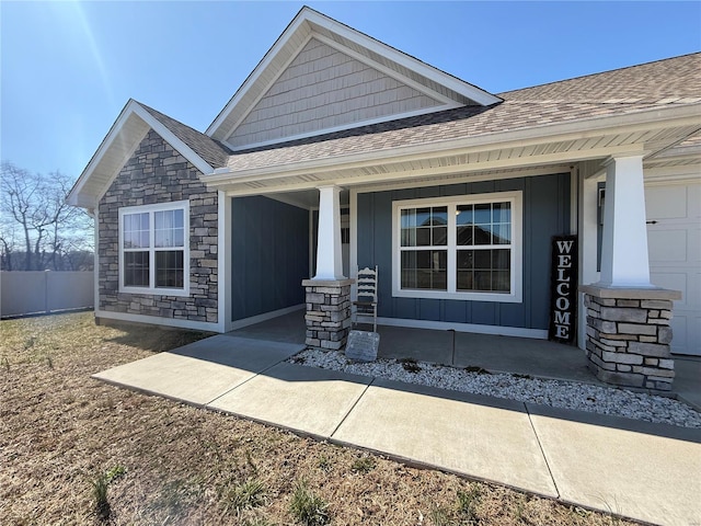 view of front of home featuring a porch, roof with shingles, stone siding, and fence