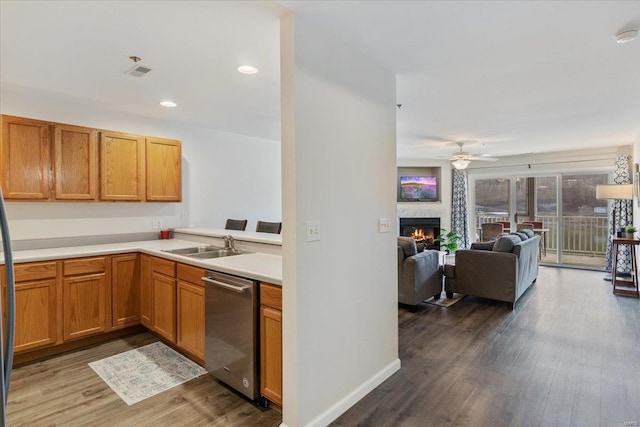 kitchen featuring stainless steel dishwasher, a ceiling fan, a sink, a warm lit fireplace, and wood finished floors