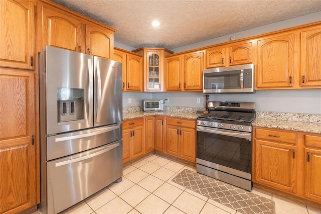 kitchen featuring appliances with stainless steel finishes, brown cabinets, glass insert cabinets, light tile patterned floors, and light stone counters