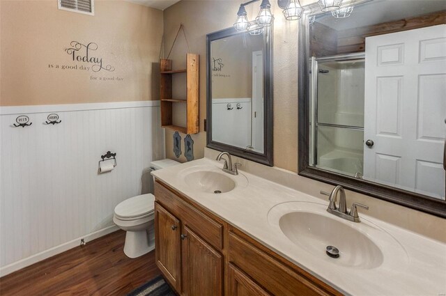 bathroom featuring a shower with door, wood finished floors, toilet, wainscoting, and double vanity