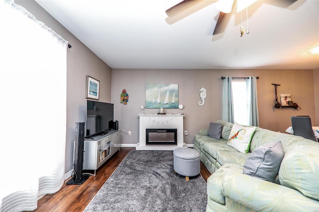 living room with dark wood finished floors, a glass covered fireplace, ceiling fan, and baseboards