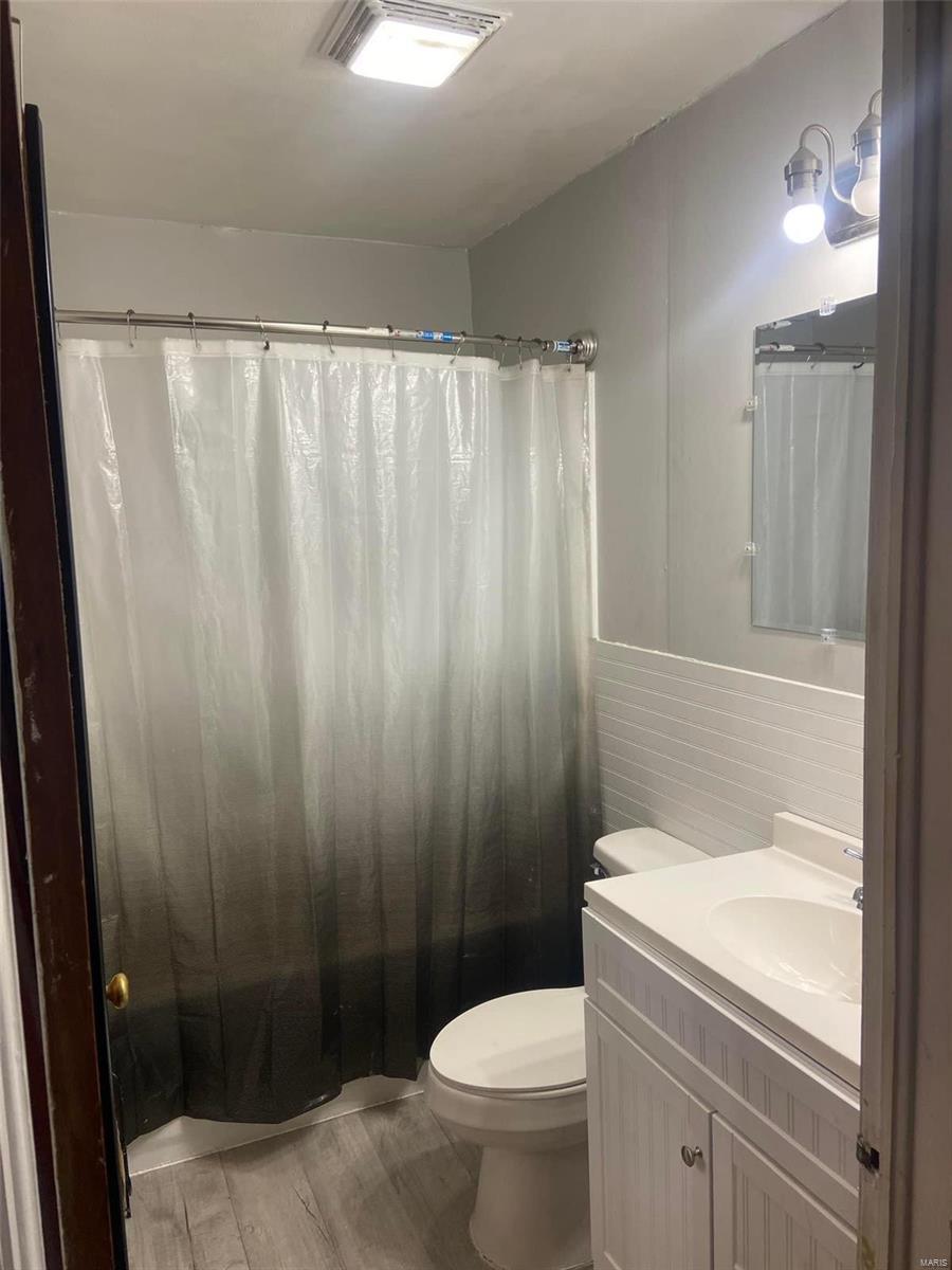 208 John R, Kennett, MO 63857 - bathroom featuring toilet, vanity, wood finished floors, a shower with curtain, and a wainscoted wall