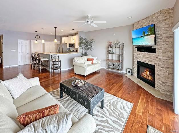 living room with ceiling fan, a stone fireplace, wood finished floors, and baseboards