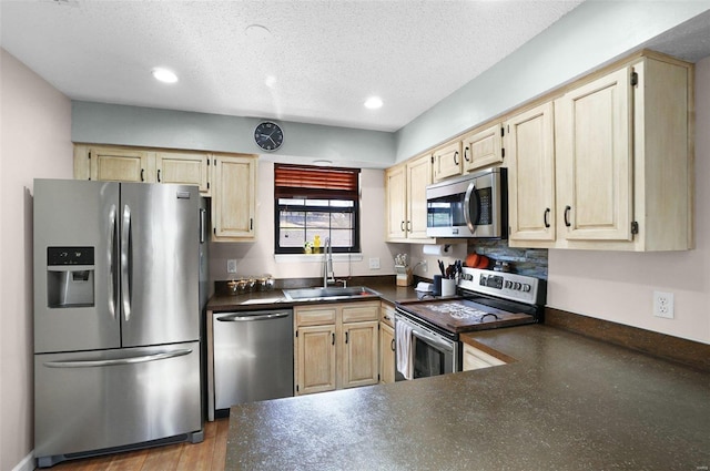 kitchen with appliances with stainless steel finishes, a sink, dark countertops, light brown cabinetry, and a textured ceiling