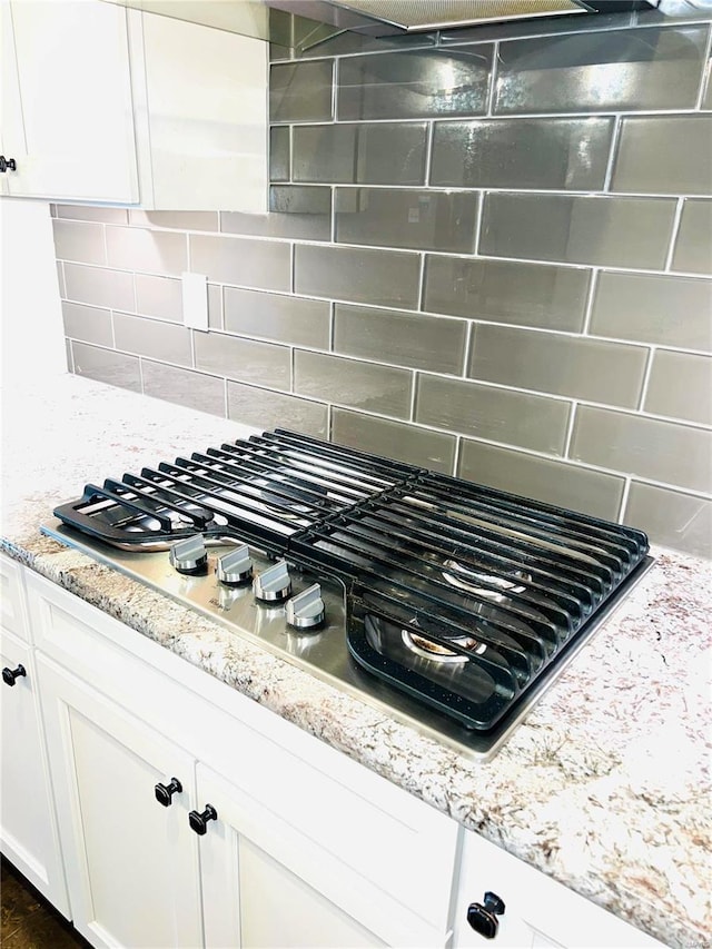 kitchen view of stainless steel gas stovetop, white cabinets, decorative backsplash, and light stone countertops