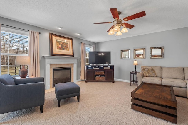 living room with light carpet, ceiling fan, baseboards, a tiled fireplace, and a textured ceiling