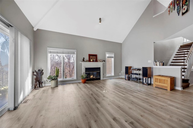 living area with high vaulted ceiling, wood finished floors, a glass covered fireplace, stairs, and healthy amount of natural light