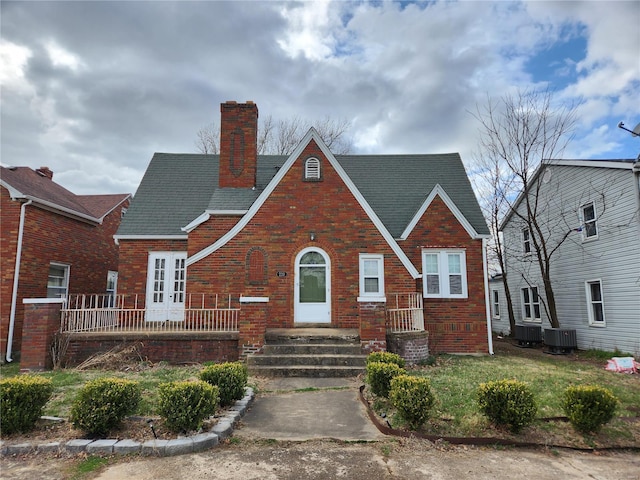tudor-style house featuring brick siding, a chimney, central AC unit, and a shingled roof