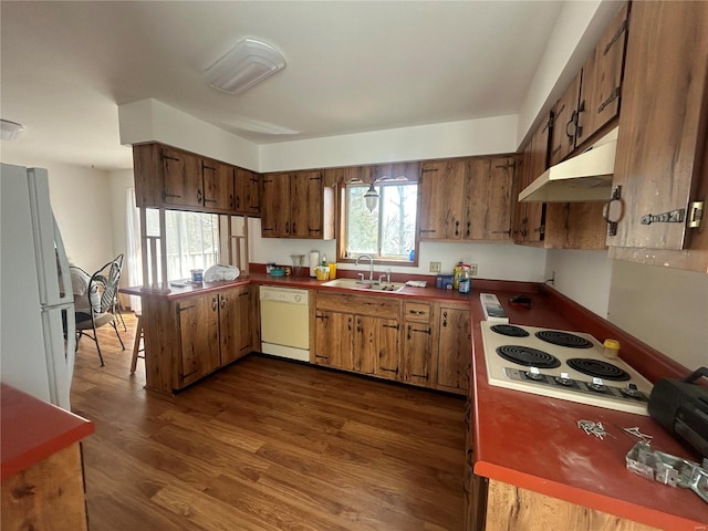 kitchen with white appliances, a sink, under cabinet range hood, dark wood finished floors, and a peninsula