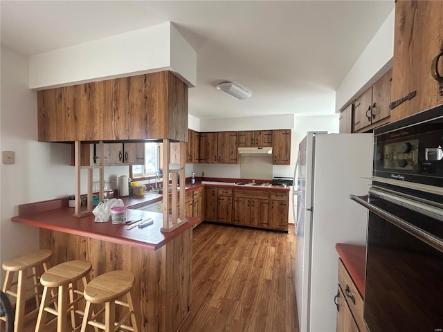 kitchen featuring white appliances, brown cabinetry, wood finished floors, a peninsula, and under cabinet range hood