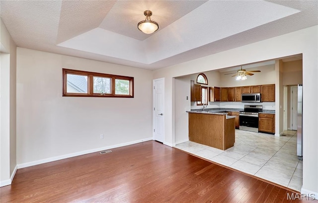 kitchen with stainless steel appliances, a raised ceiling, light wood-type flooring, brown cabinetry, and a peninsula