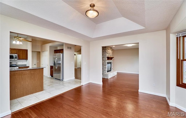 unfurnished living room with light wood-type flooring, a ceiling fan, a stone fireplace, a tray ceiling, and baseboards