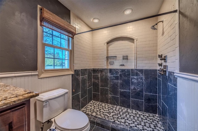 full bathroom featuring a textured ceiling, a wainscoted wall, tiled shower, toilet, and vanity