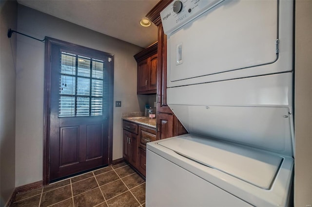clothes washing area featuring stacked washer / drying machine, baseboards, dark tile patterned flooring, and cabinet space