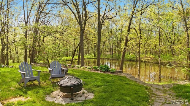 view of green lawn with a fire pit, a forest view, and a water view
