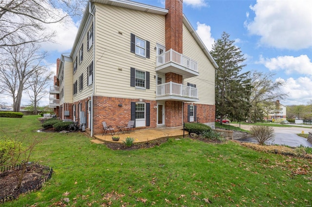 back of property featuring a balcony, a chimney, brick siding, a lawn, and a patio